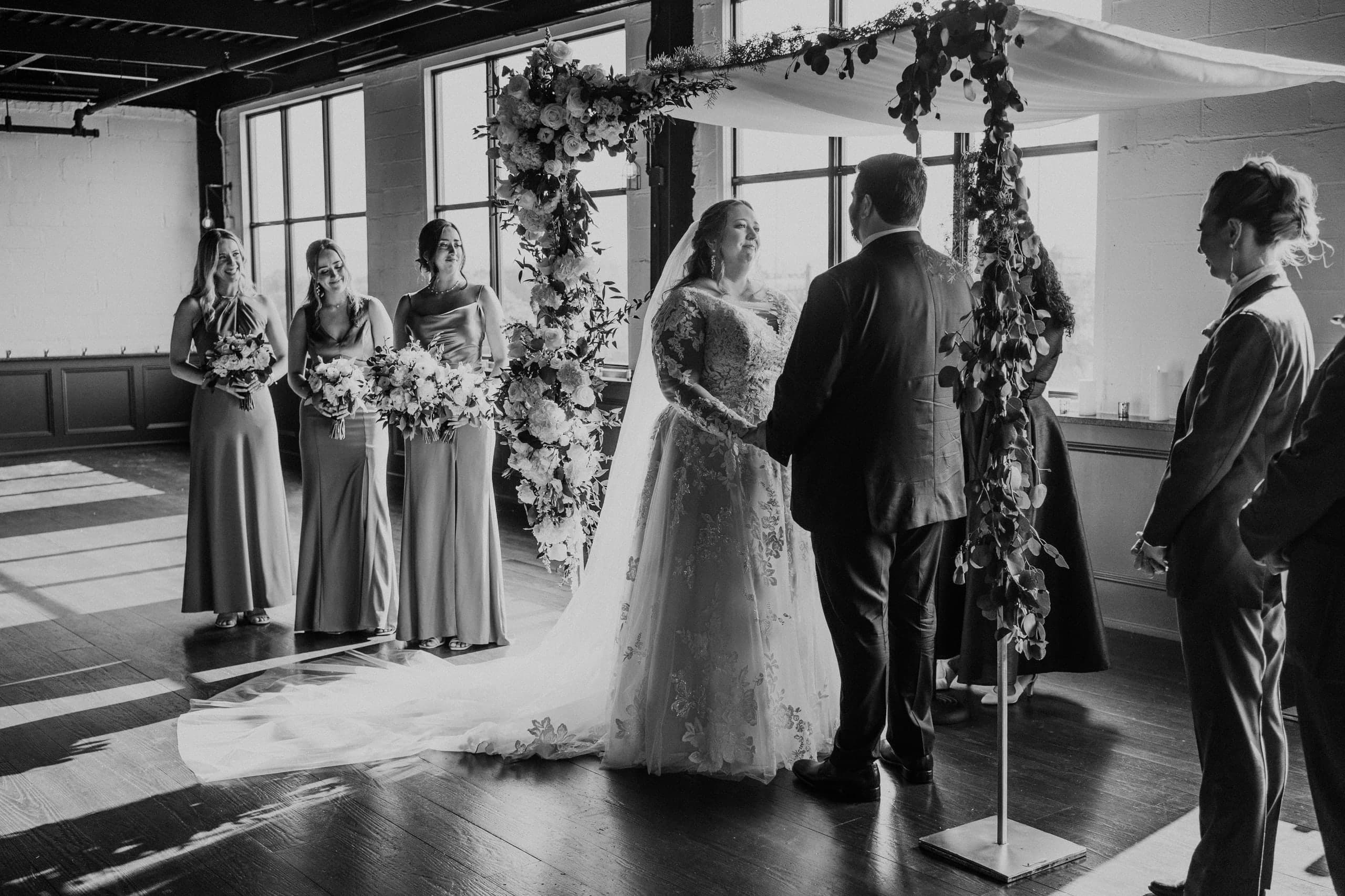 Vows beneath a floral canopy in a loft flooded with window light, black and white