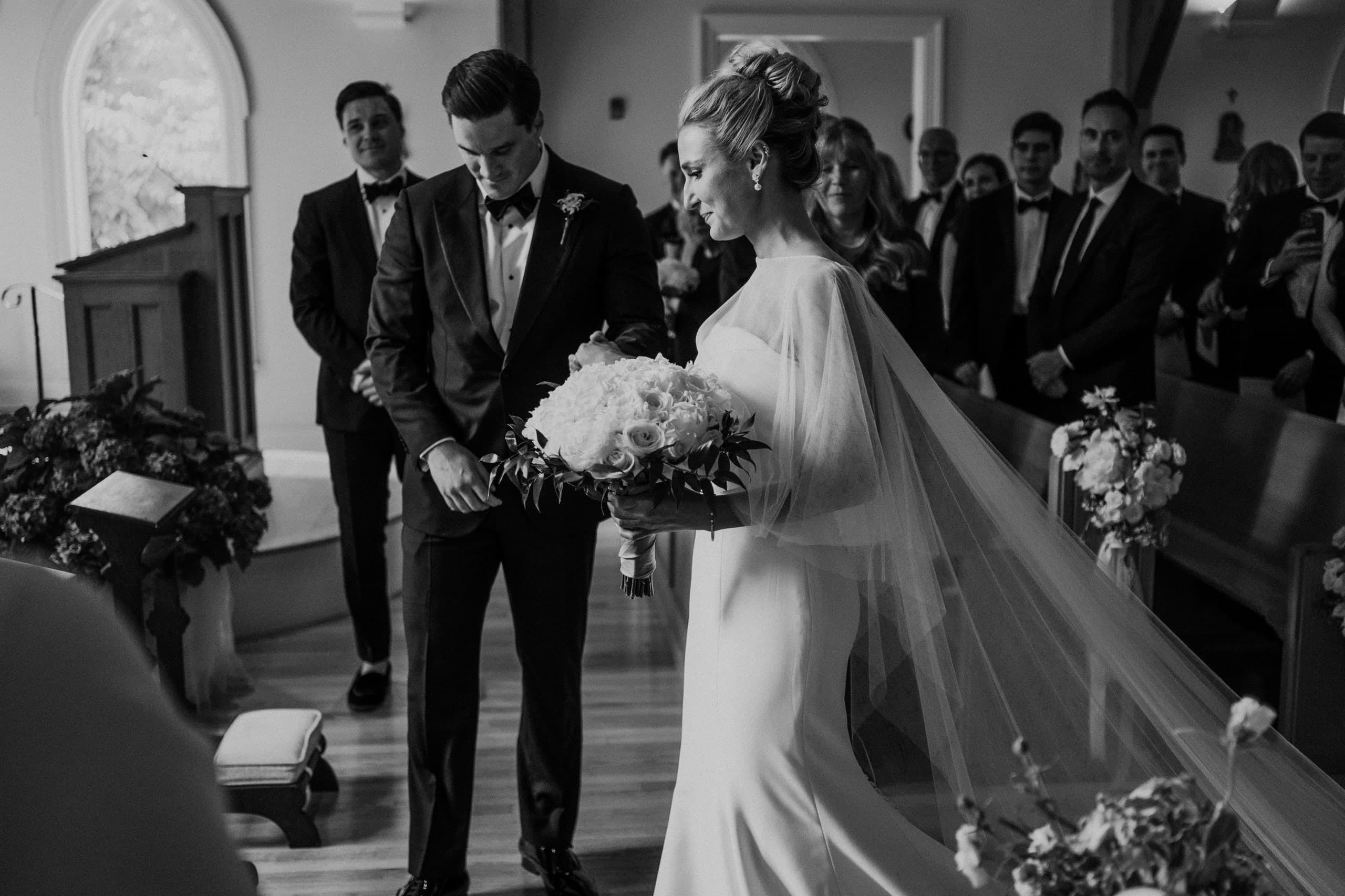 The bride's sheer cape trailing as the couple meets at the chapel altar, black and white
