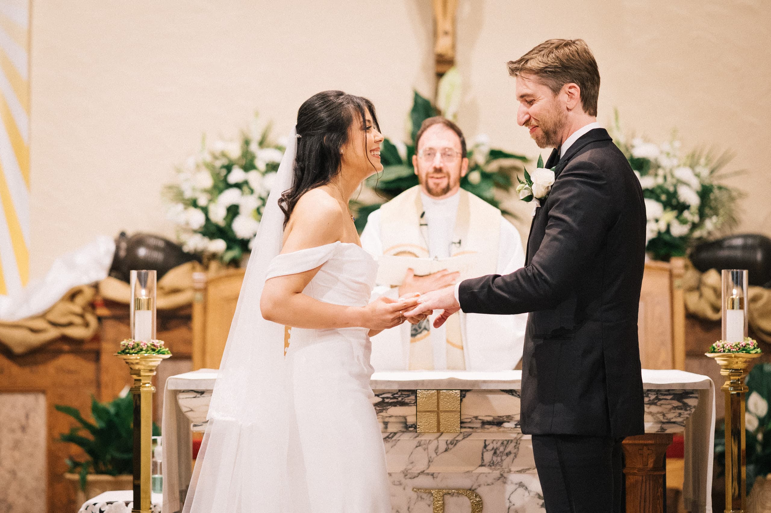 Exchanging rings at a flower-banked altar, the priest smiling between them