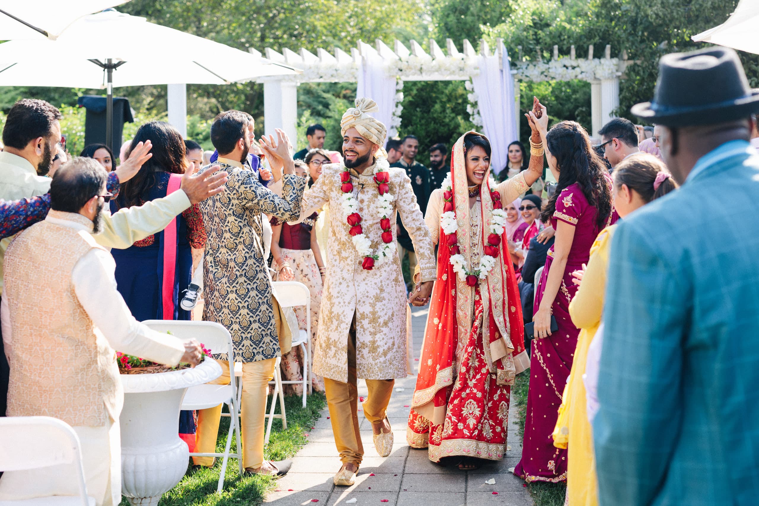 The couple in red and gold showered with cheers down a sunlit garden aisle