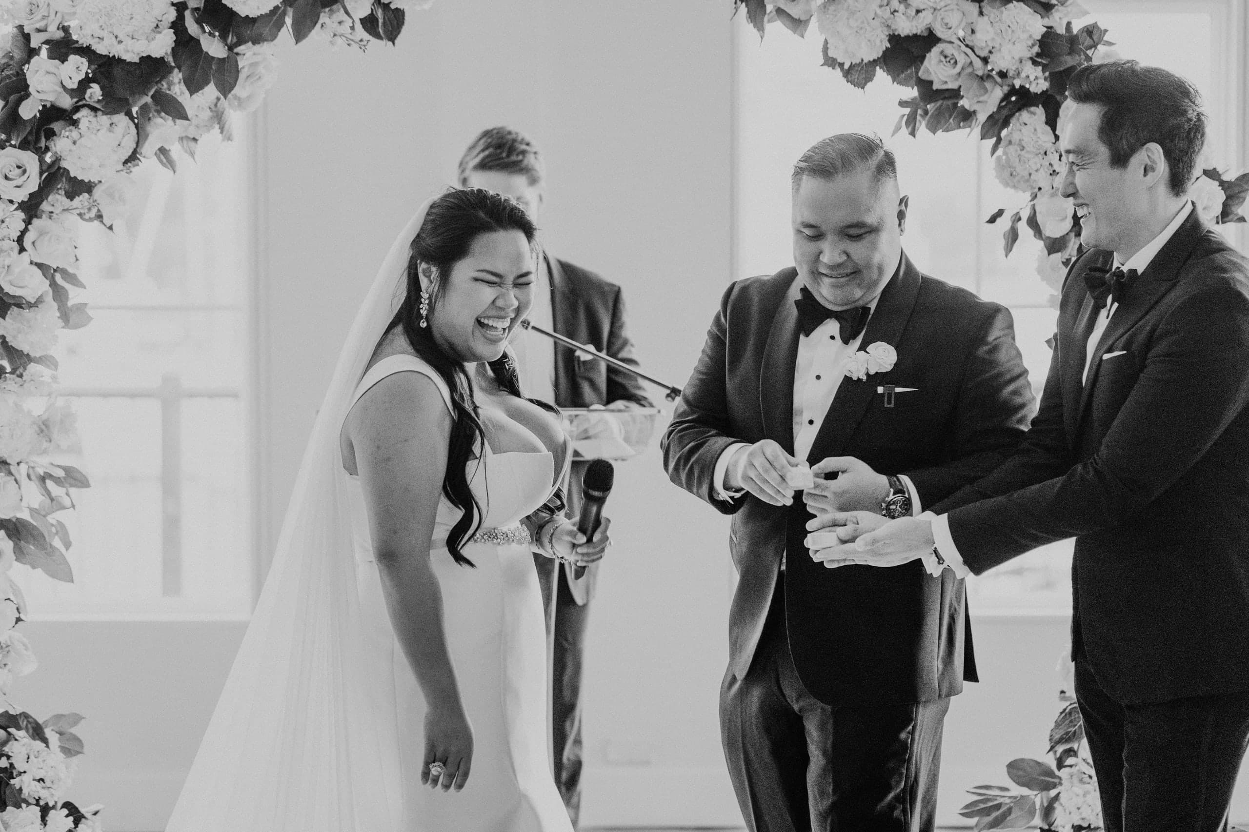 The bride laughing beneath a rose-covered arch as the rings come out, black and white