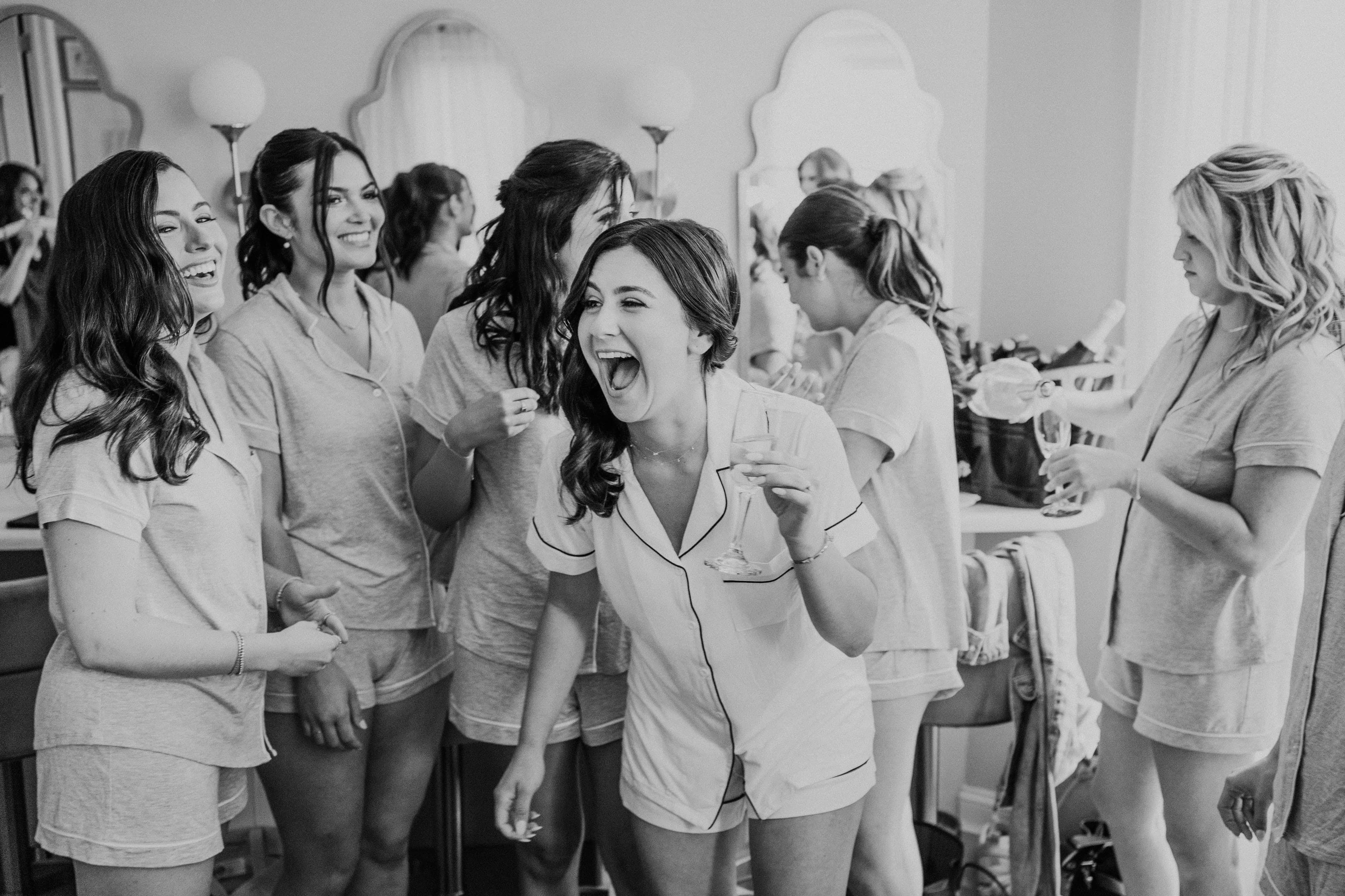 Bridesmaids in matching pajamas erupting in laughter around the vanity, black and white