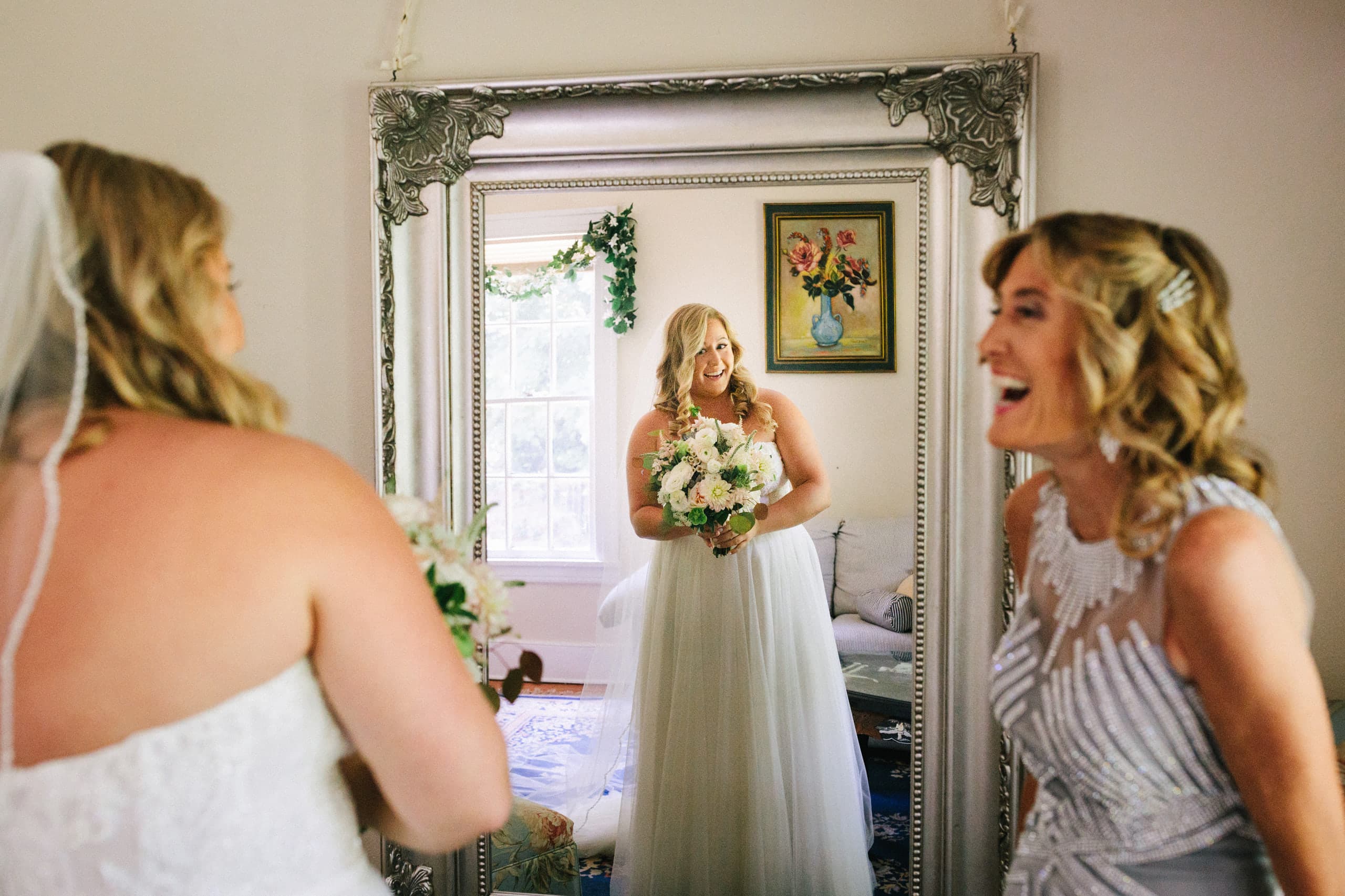Bride with her bouquet framed in a tall silver mirror beside a laughing bridesmaid