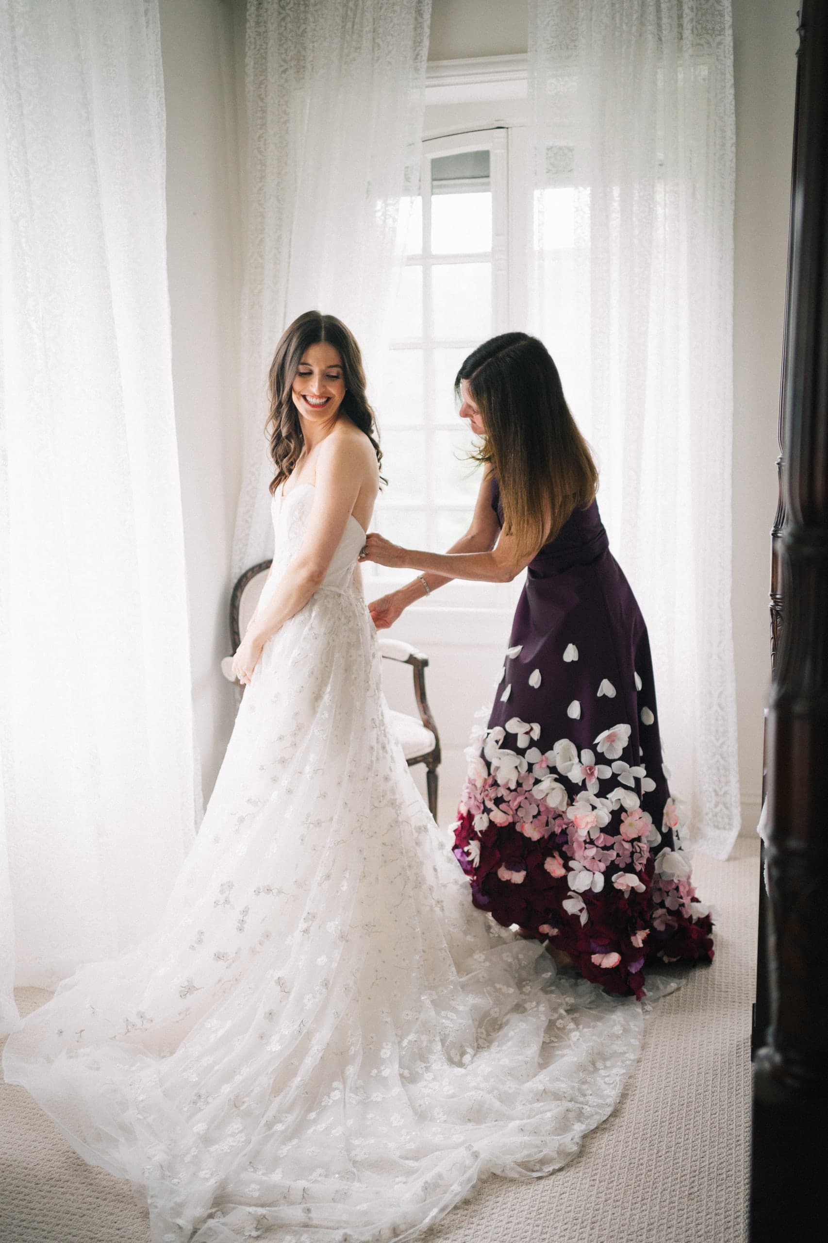 Bride glancing over her shoulder as her mother buttons her gown by a sheer-curtained window