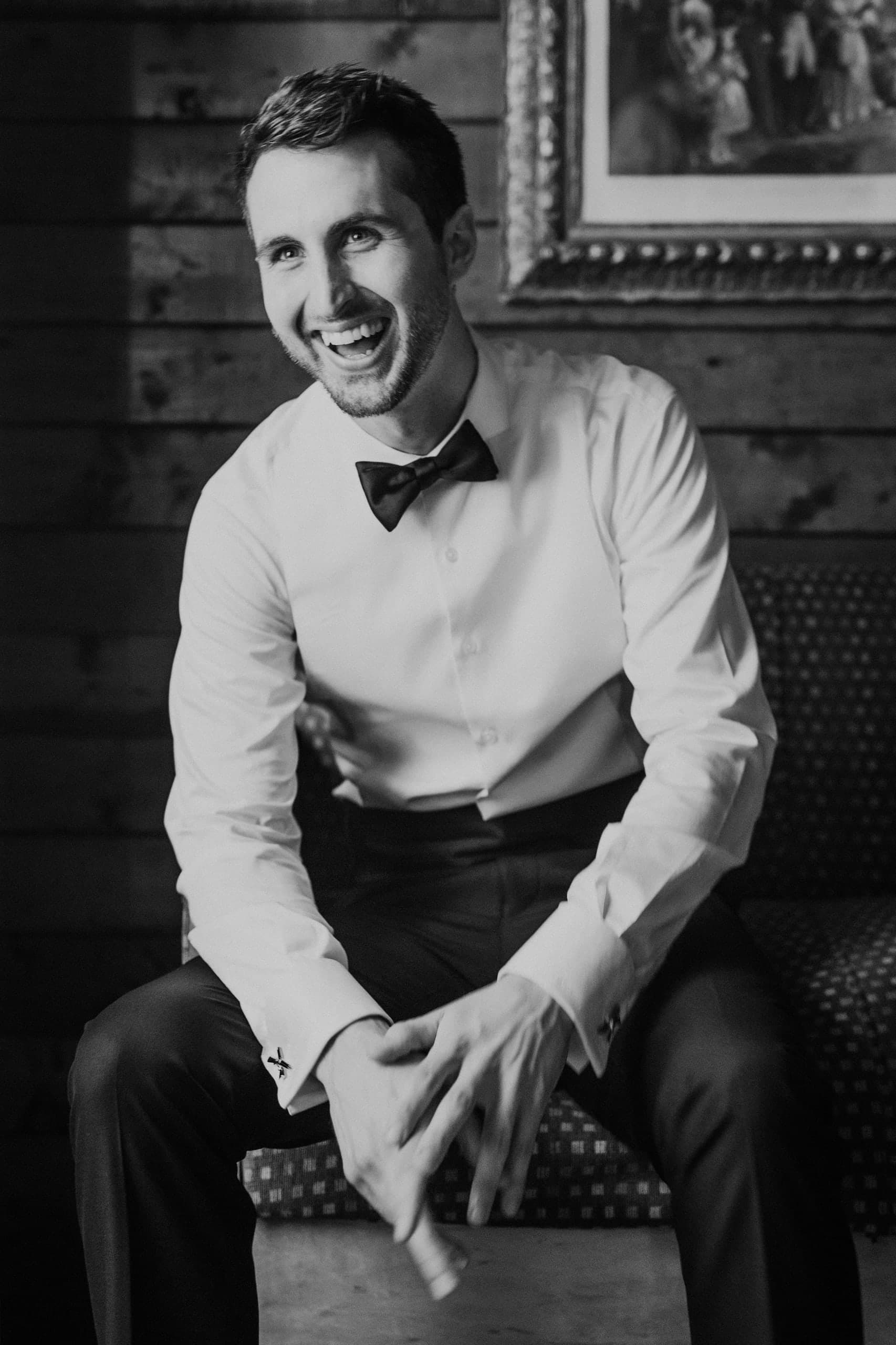 Groom in shirtsleeves and bow tie laughing on a bench against a wood-paneled wall, black and white