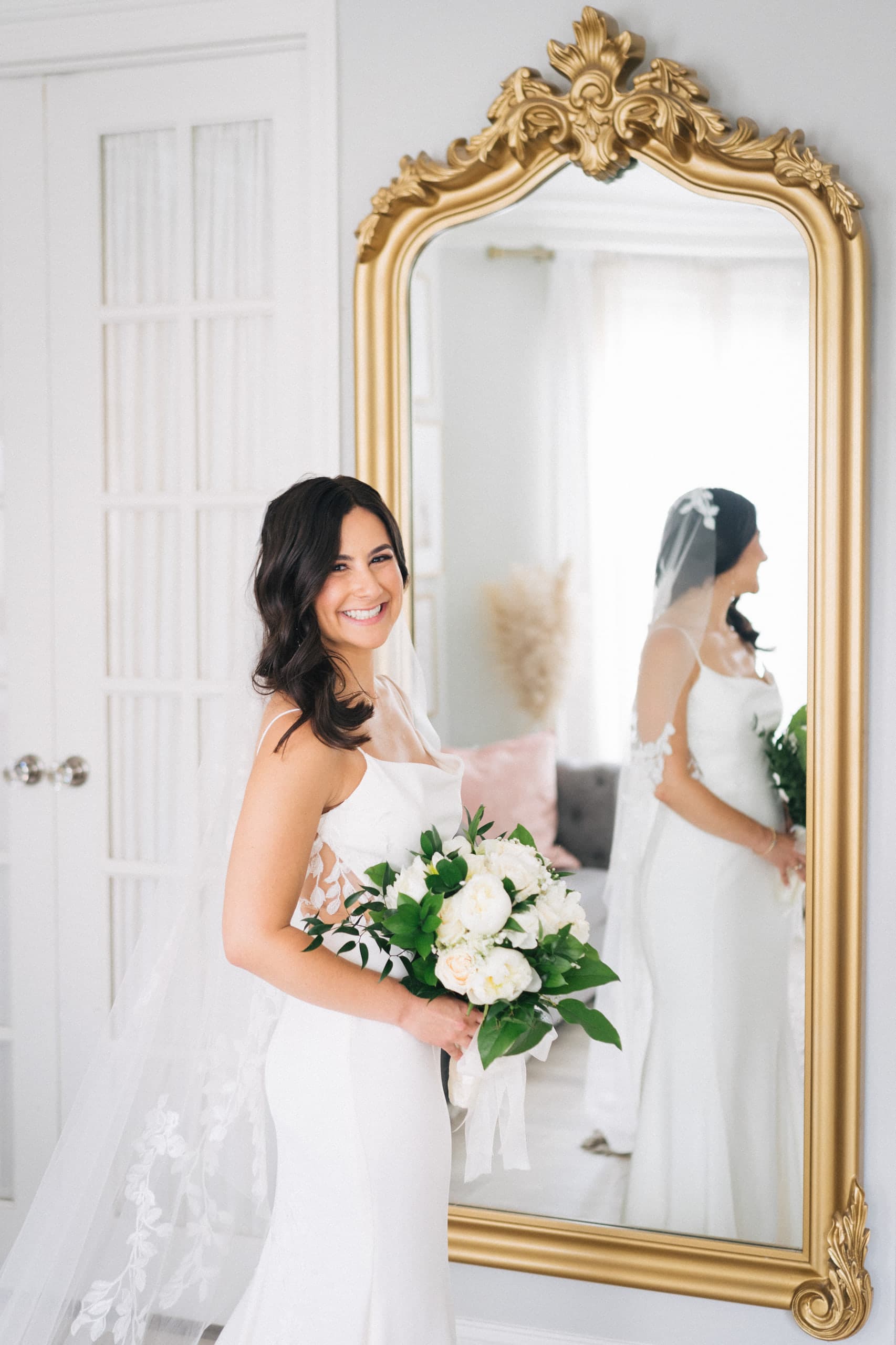 Smiling bride with a white bouquet beside an ornate gold mirror catching her veiled reflection