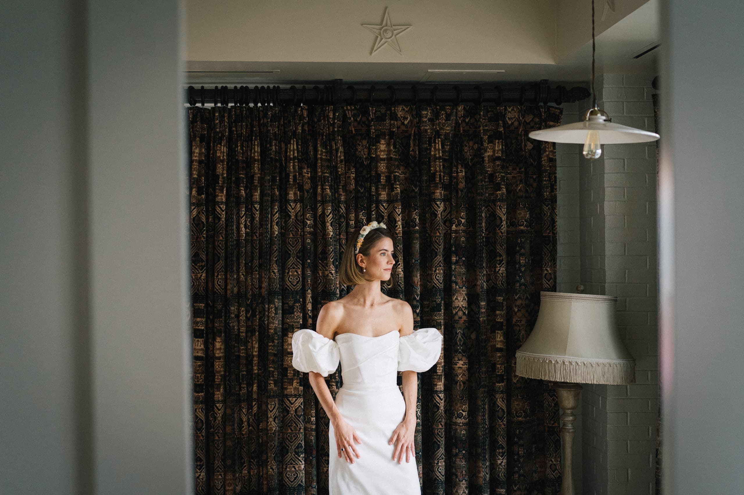 Bride in a puff-sleeve gown framed in a doorway before a dark patterned curtain and vintage lamps