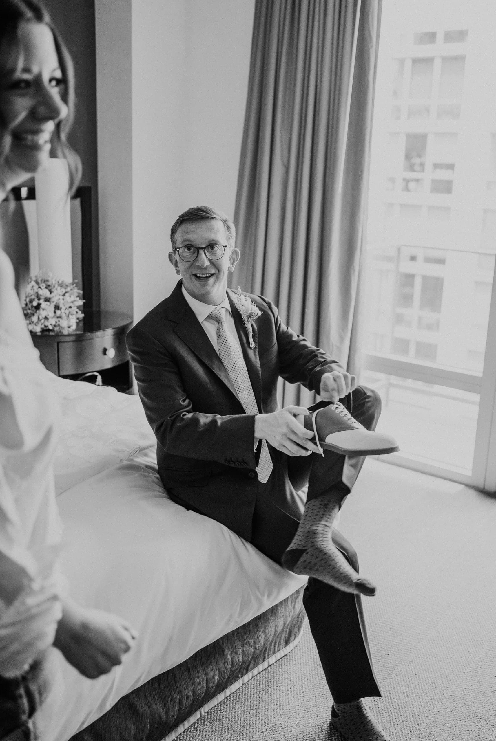 Grinning groom seated on the bed pulling on his shoes in window light, black and white