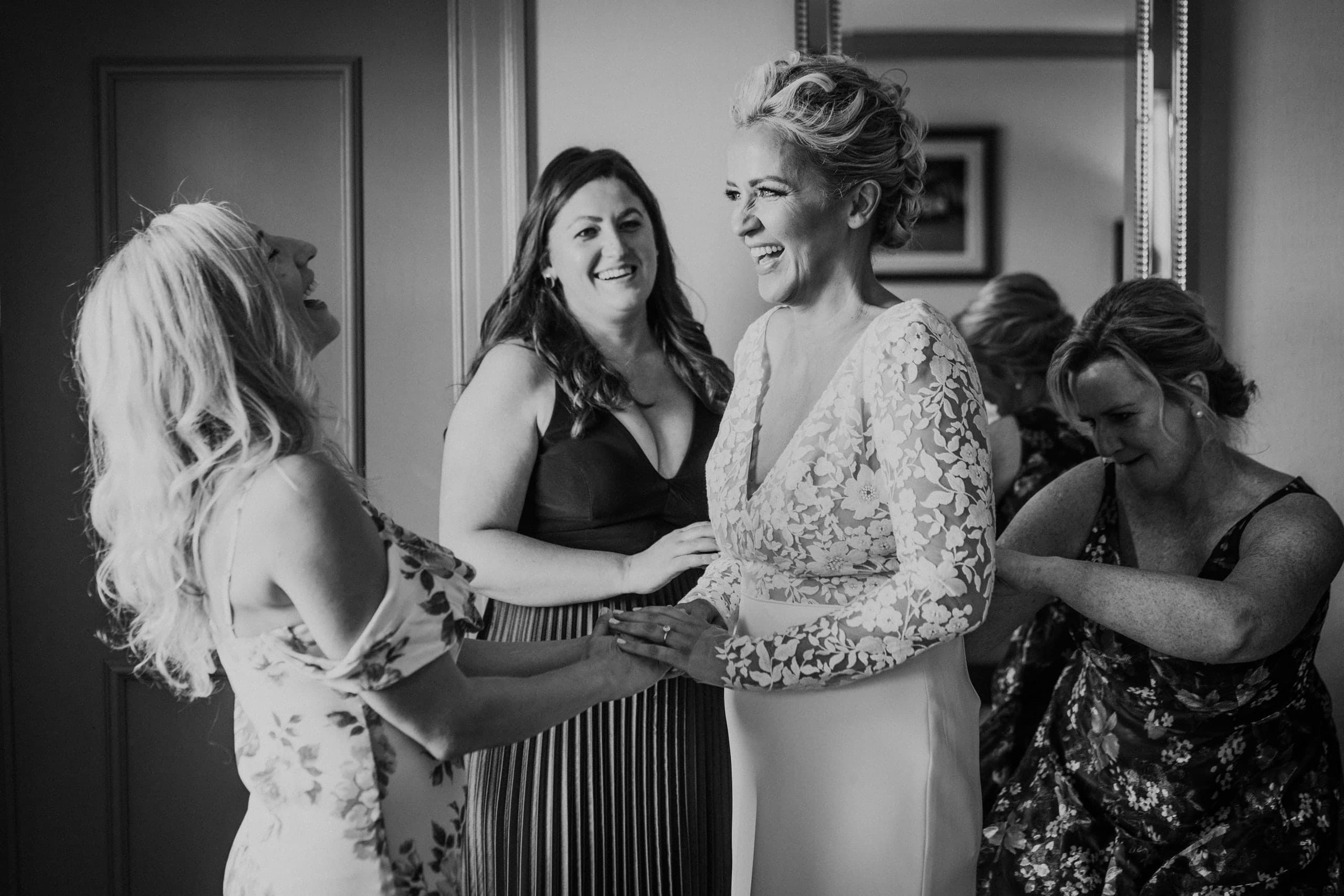 Bride in lace sleeves laughing with her bridesmaids as one fastens her gown, black and white