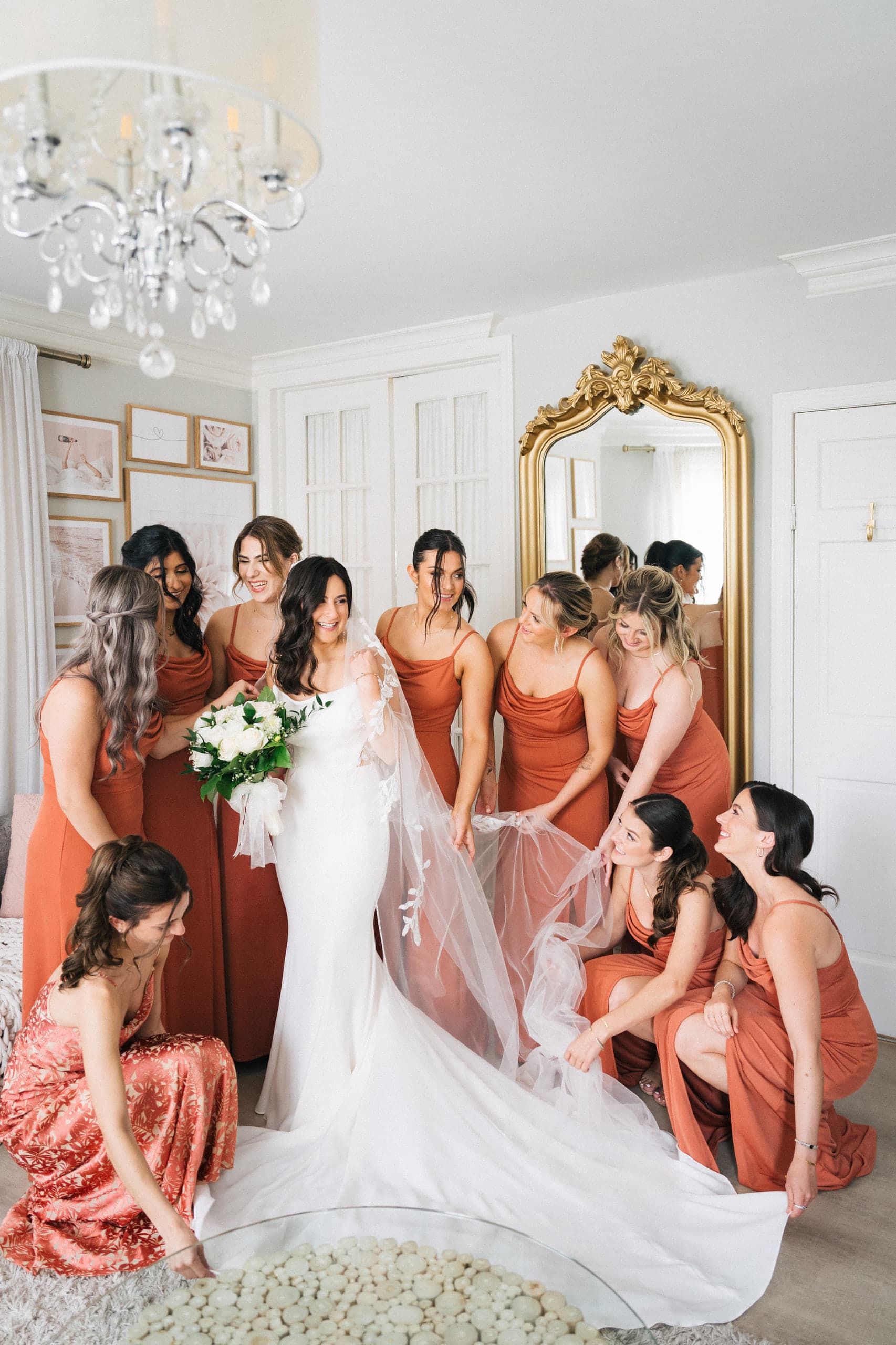 Bridesmaids in rust gowns fanning the bride's veil beneath a chandelier and gold mirror