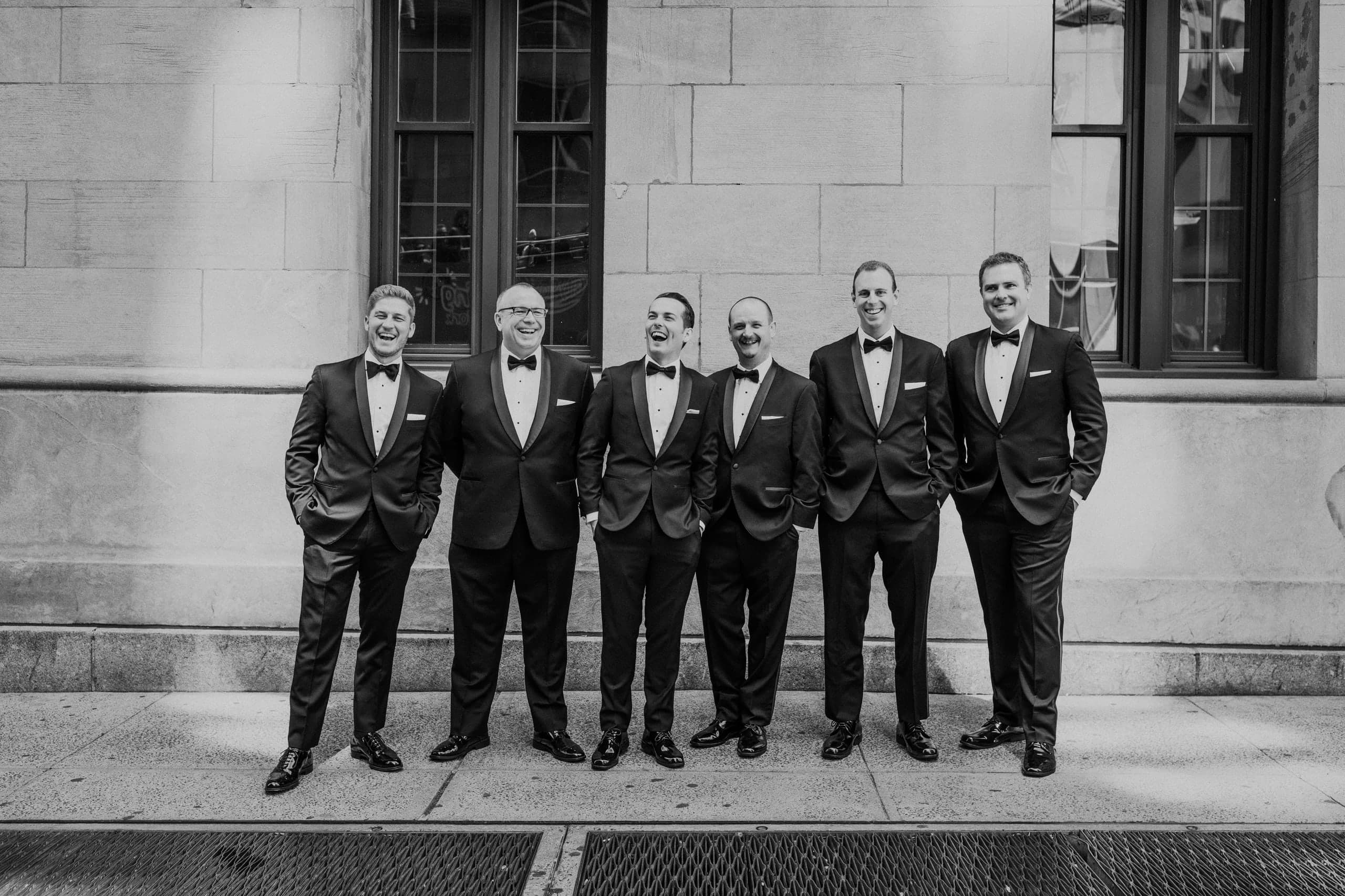 Six groomsmen in tuxedos lined up laughing along a stone facade, black and white