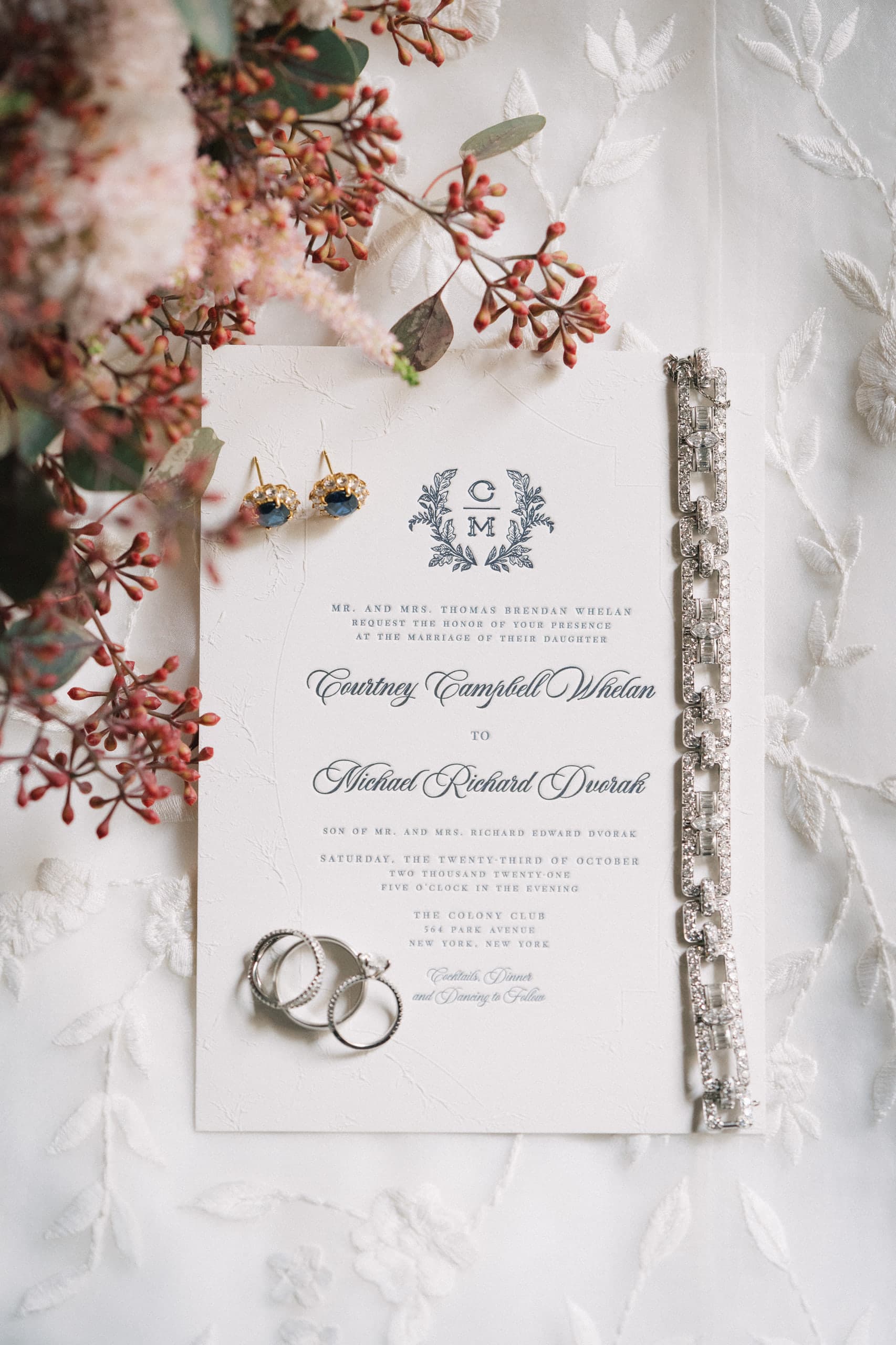 Wedding invitation flat lay with rings, sapphire earrings, and a crystal bracelet amid blooms