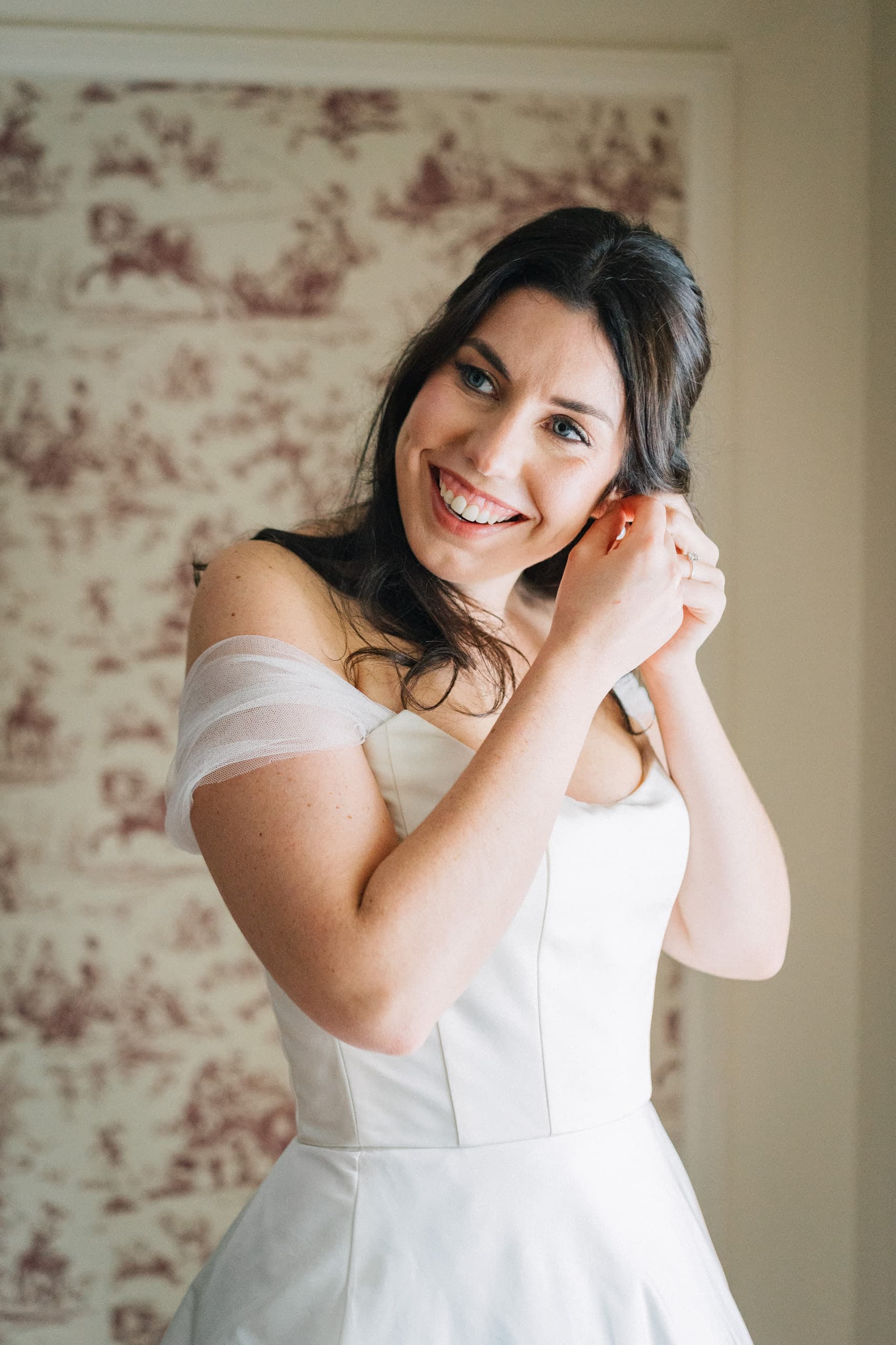 Bride putting on her earring before red toile wallpaper, smiling toward the window