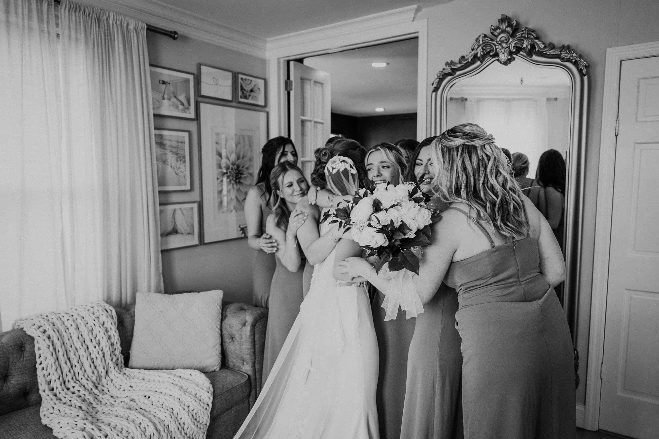 Bridesmaids wrapping the bride and her bouquet in a group hug by the mirror, black and white