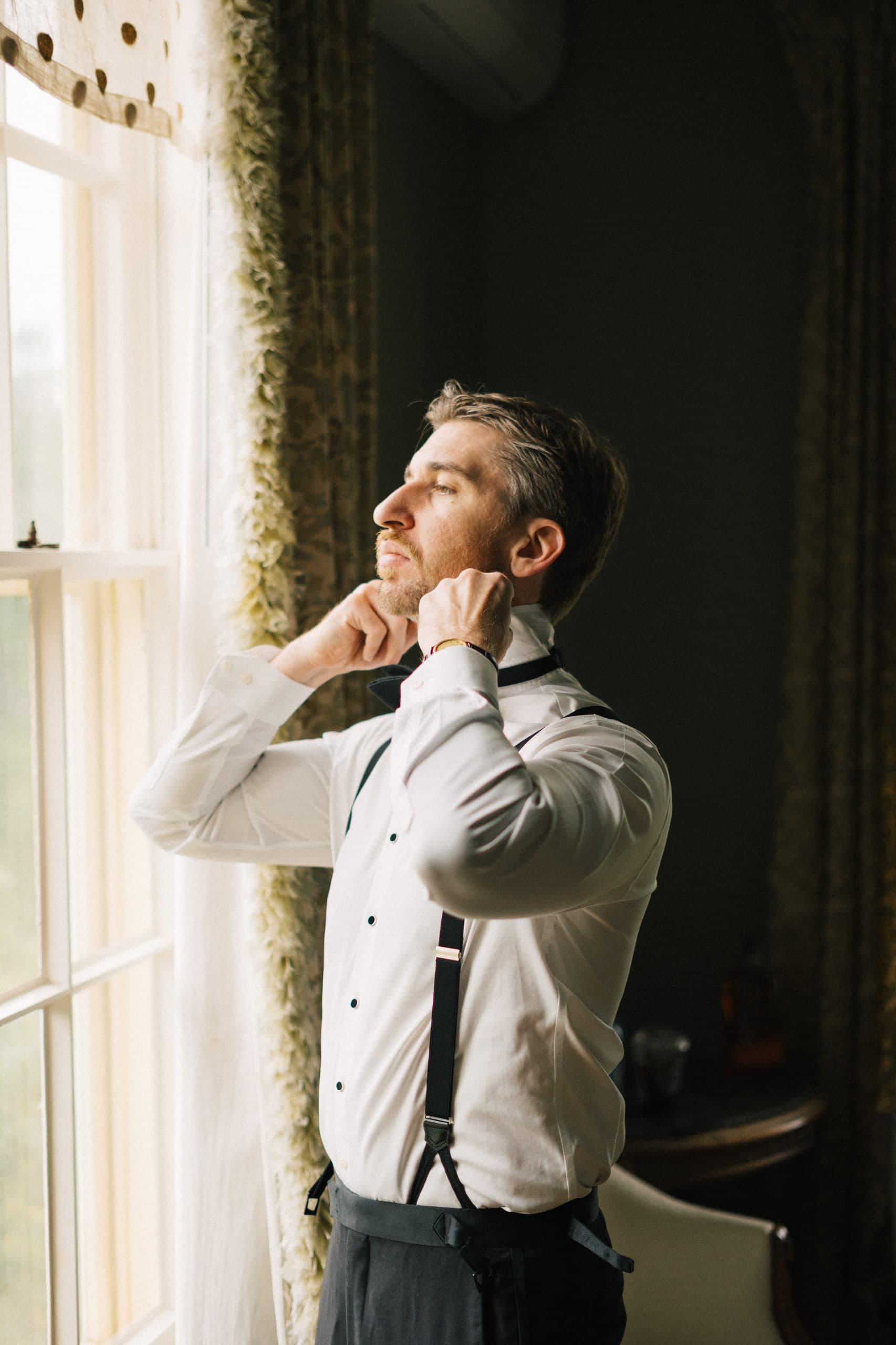 Groom in suspenders adjusting his bow tie in soft window light