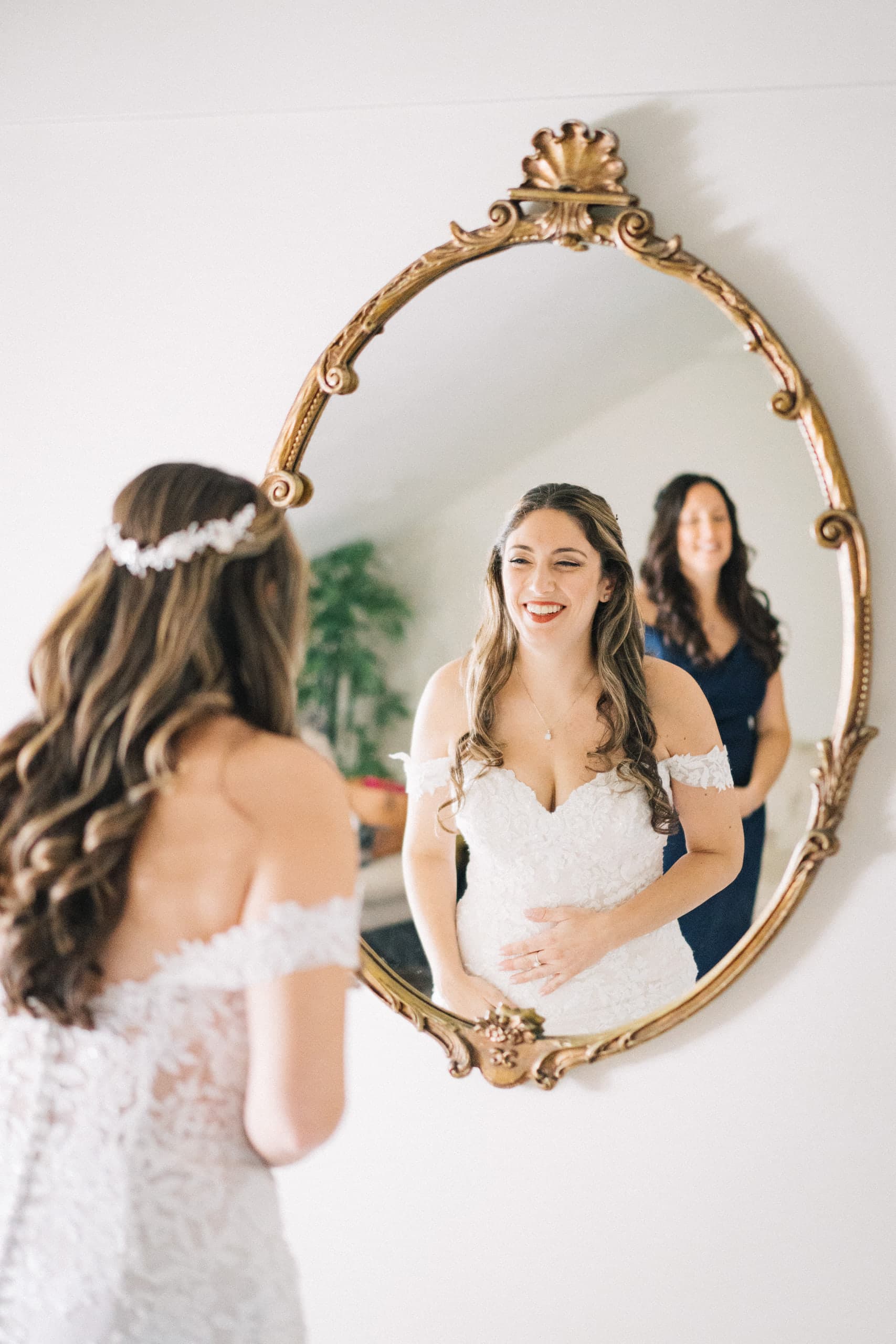 Bride smiling at her reflection in an oval gilt mirror, her mother watching behind