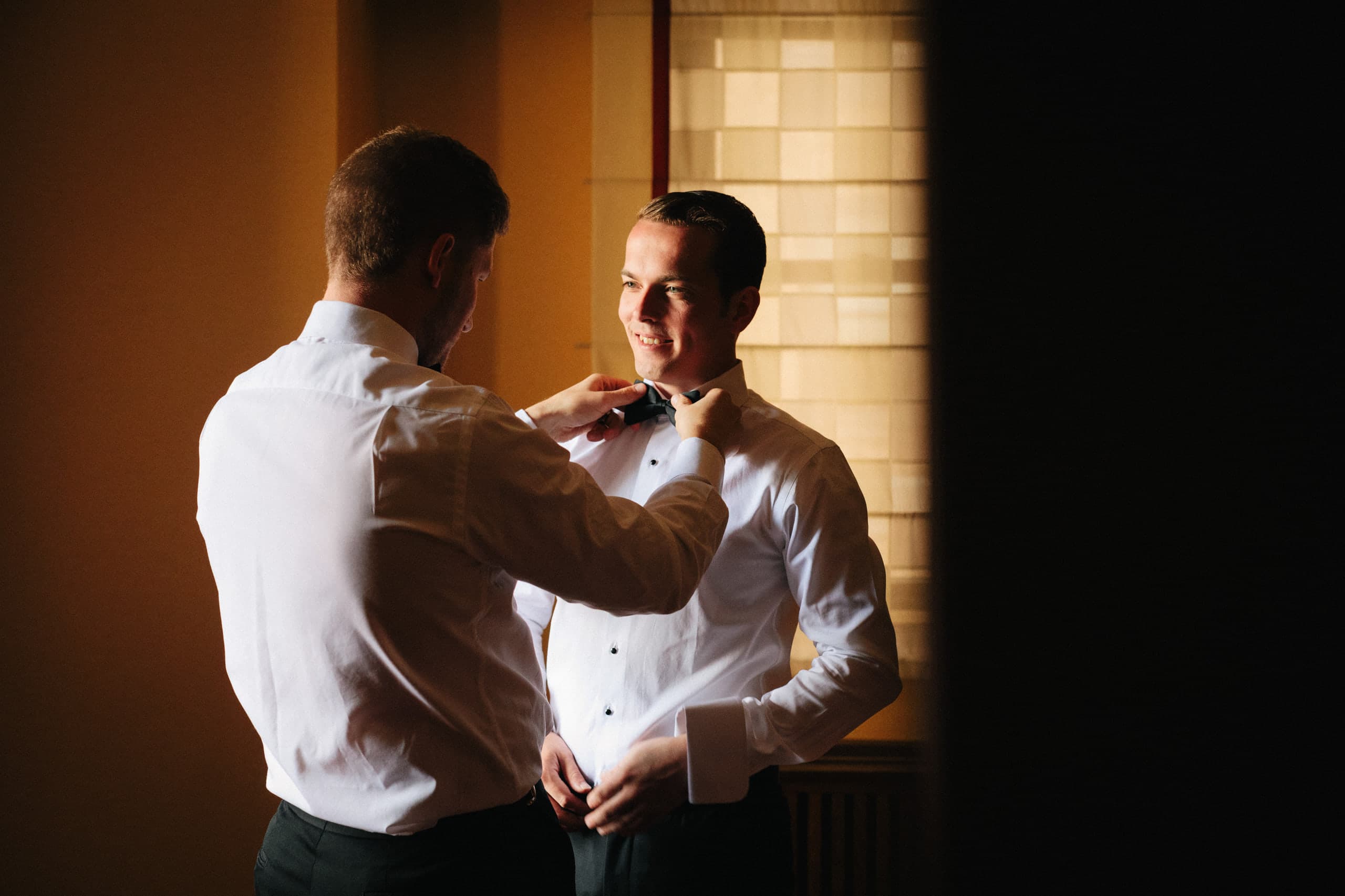 Groomsman straightening the groom's bow tie in warm amber window light