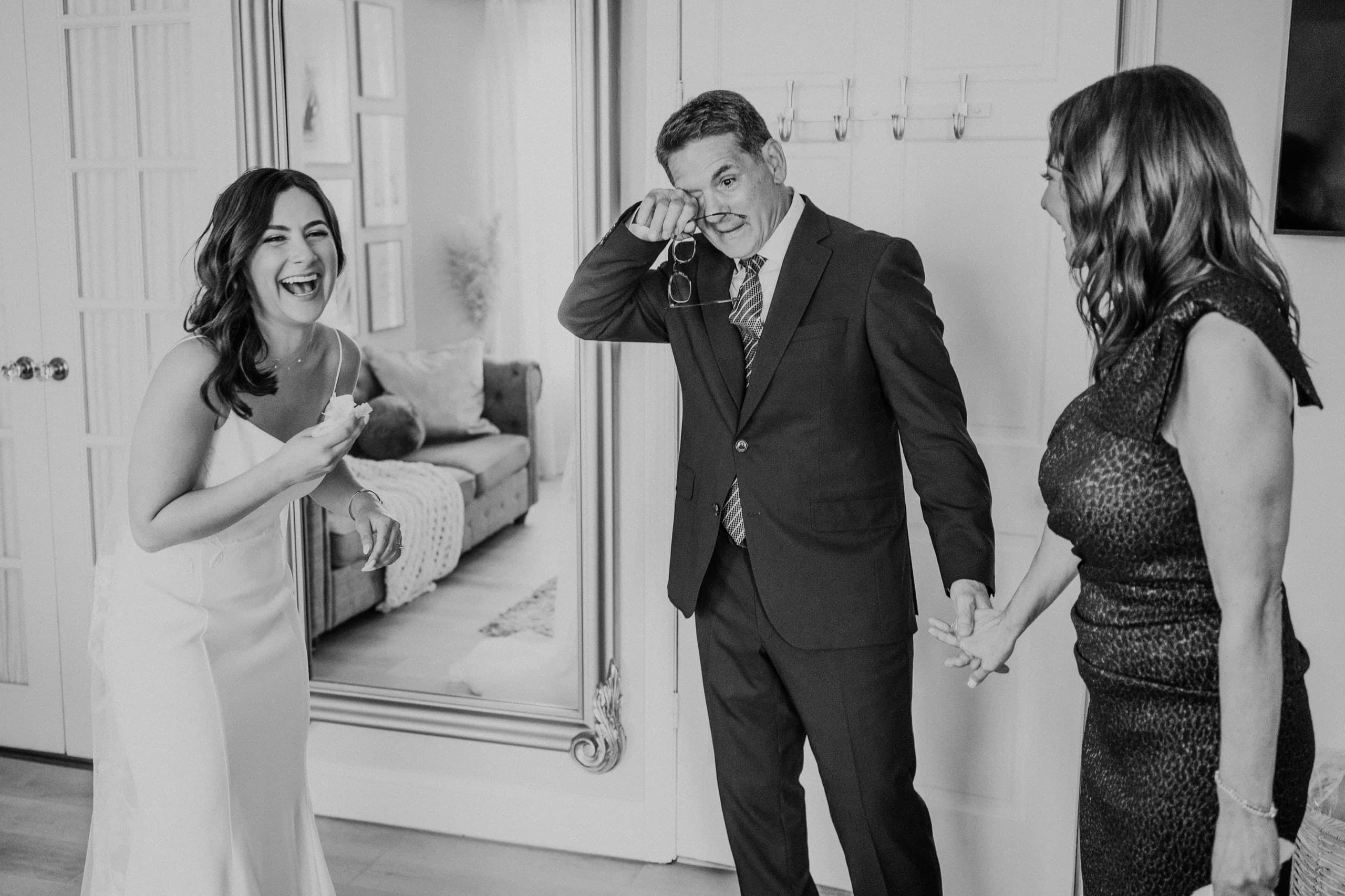 Father wiping away a tear as the laughing bride shows him her gown, black and white