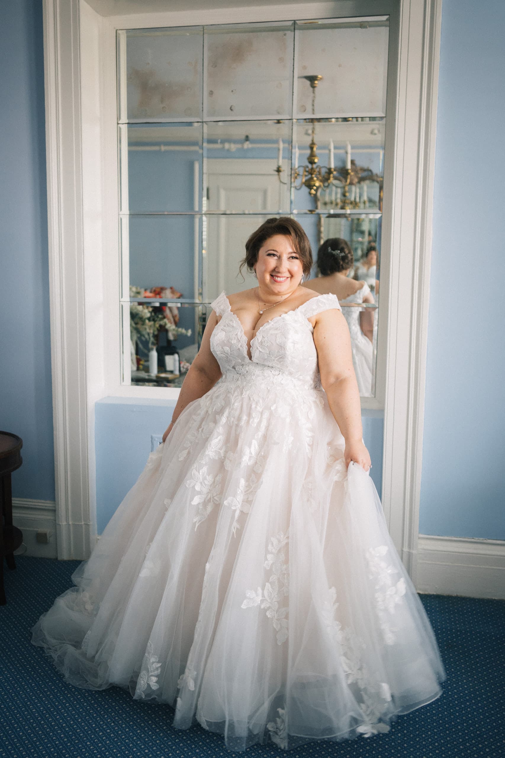 Bride in a lace ball gown smiling before a mirrored wall in a pale blue room