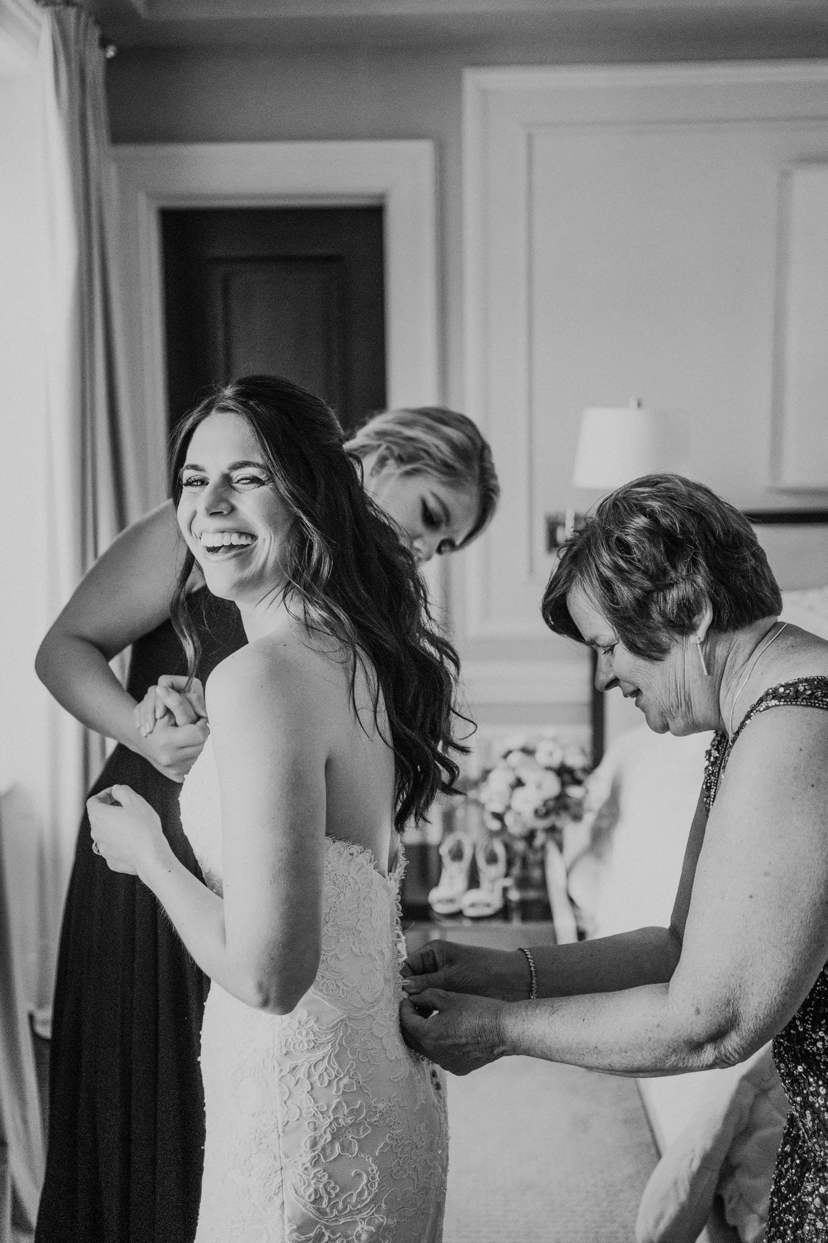Bride laughing over her shoulder as her mother buttons the back of her gown, black and white