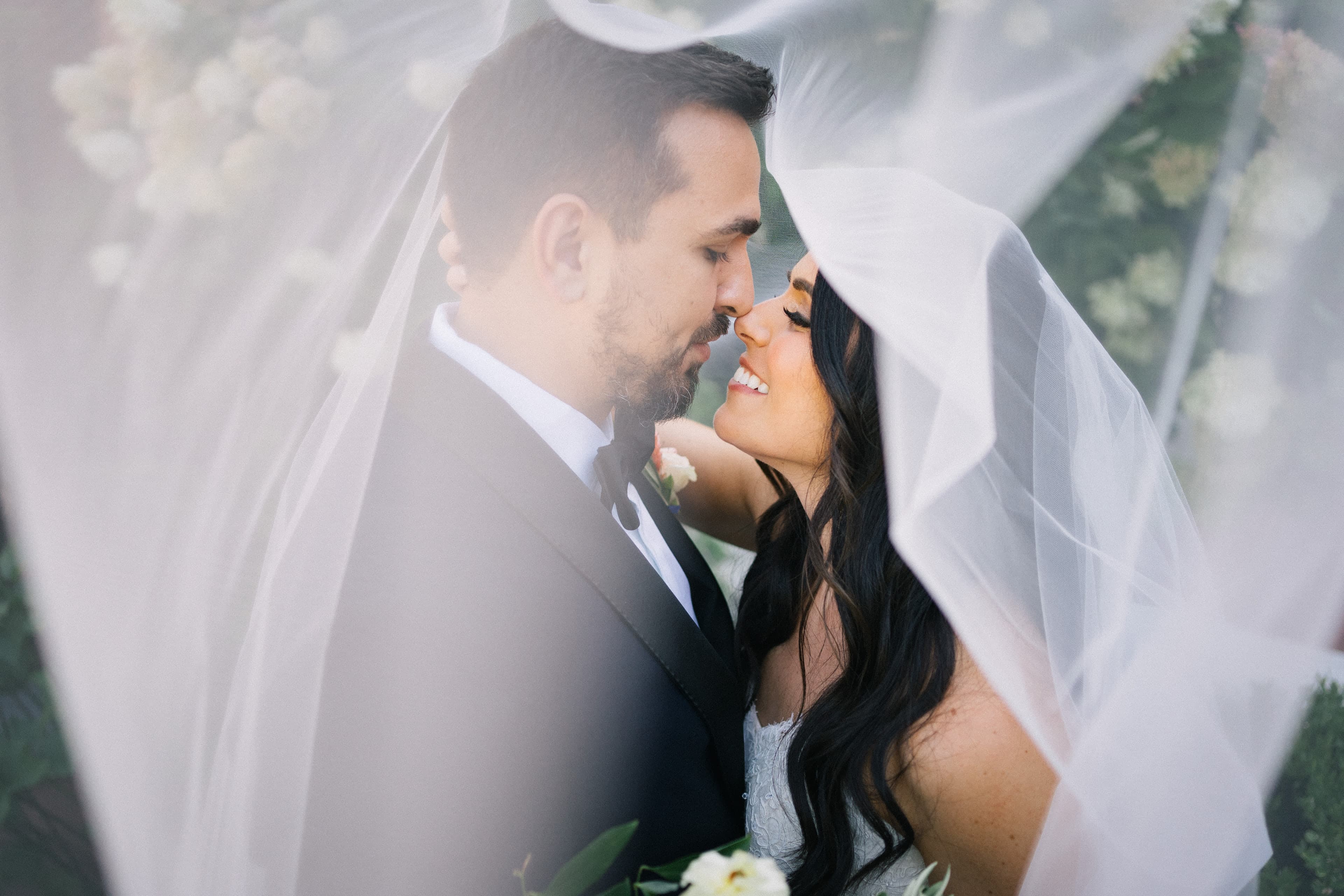A bride and groom smile nose to nose beneath her billowing sheer veil, surrounded by white blooms