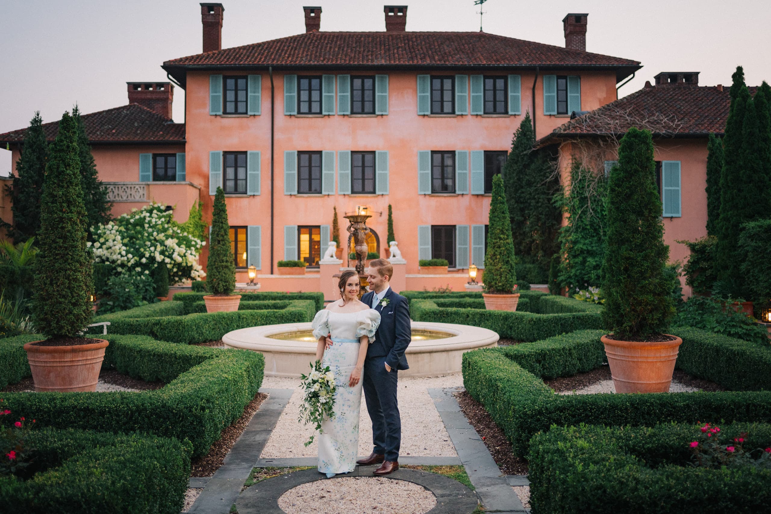 Standing at the fountain in the parterre garden of a rose-pink villa at dusk