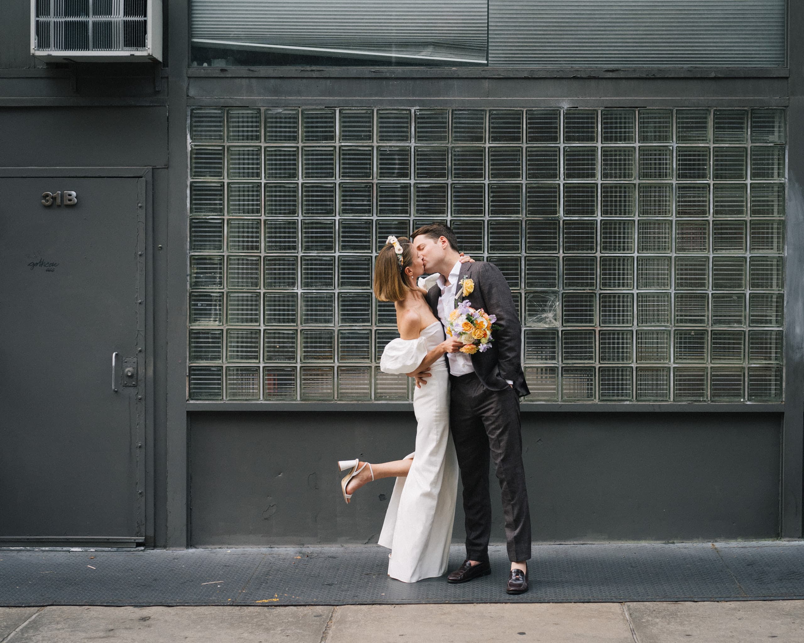 A kiss on the sidewalk against a glass-block wall, one heel kicked up