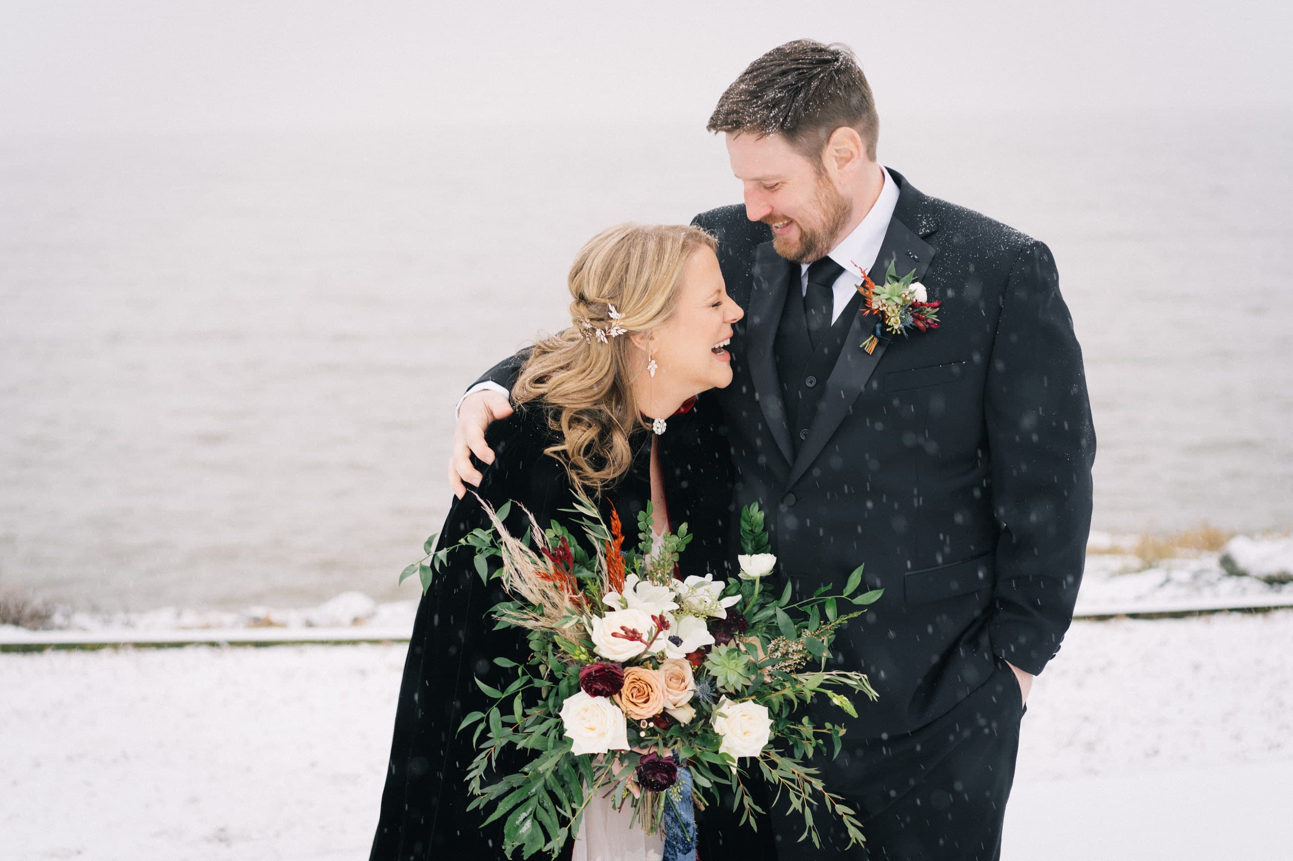 Laughing under falling snow at the water's edge, bouquet in hand
